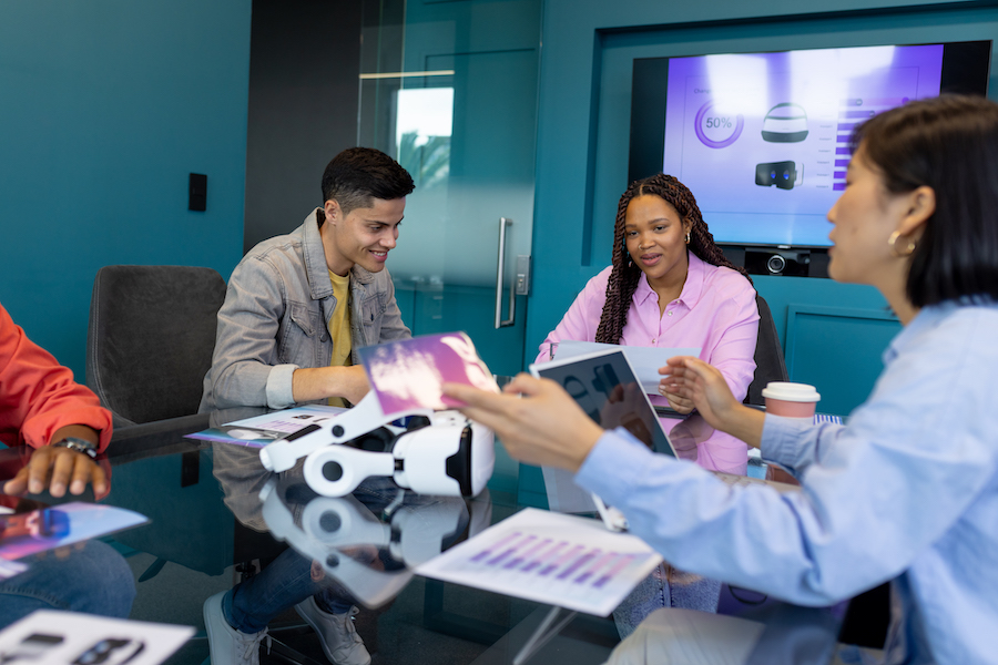 Colleagues from diverse backgrounds discuss VR tech in a modern business office. They are surrounded by VR headsets and charts, with a presentation screen in background, unaltered